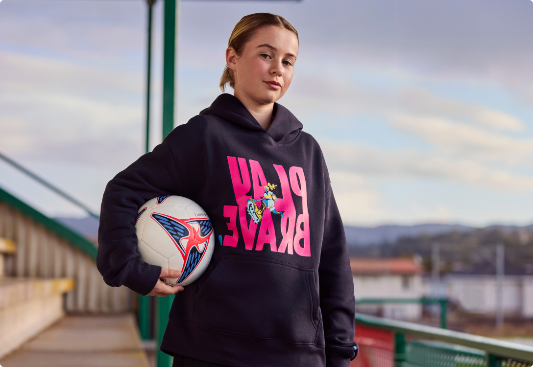SupportHER Merch - Reverse merch Young girl wearing the Play brave hoodie holding onto a football